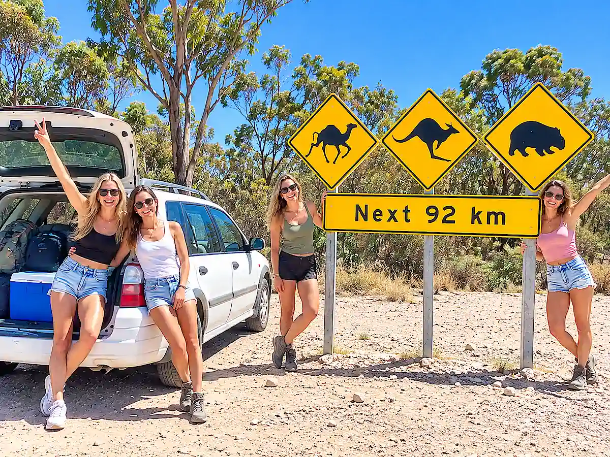 Vier Reisende mit einem Kombi auf einem Australien-Roadtrip neben Straßenschildern für Kängurus, Wombats und Kamele
