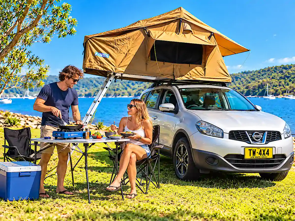 SUV Camper kaufen – Paar kocht gemeinsam am See neben ihrem Nissan SUV mit geöffnetem Dachzelt in Australien 
